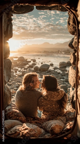 A couple sits intimately within a stone archway overlooking a rocky beach at sunset, with a cloudy sky and distant mountains in.