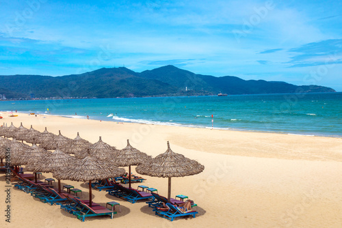 Panoramic view of Da Nang beach with thatched umbrellas, sun loungers, swimmers, and mountains along the turquoise coastline