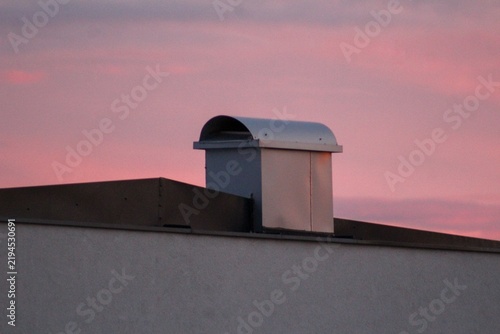 Modern metal rooftop ventilation chimney on a contemporary building under a beautiful pink sunset sky. Minimalist architectural composition with soft evening colors, clean lines, and ample copy space.
