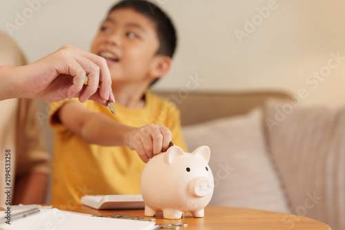 Konstfotografi Asian boy putting coin in piggy bank guided by parent for savings habit and educ