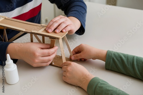 Children hands building cardboard bridge model with popsicle sticks and glue. Siblings repairing school craft project on table. Hands-on STEM education concept