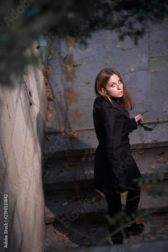 Fotografía A young woman in a black coat looks up at the camera while standing on stone steps strewn with autumn leaves, holding sunglasses; moody, contemplative urban fashion portrait in soft directional light