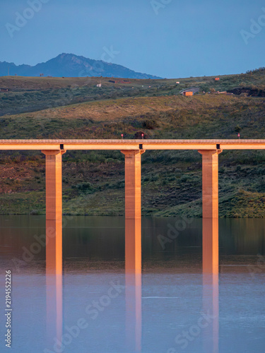 Fotografie A vertical photo capturing the tall concrete pillars of a modern bridge reflecting cleanly in the mirror-like surface of a tranquil lake during twilight