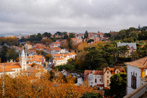 Panoramic view of Sintra town with red tile roofs, autumn trees and church tower under dramatic cloudy sky in Portugal.