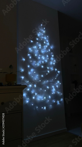 White string lights arranged in the shape of a Christmas tree glow on a wall in a dark room