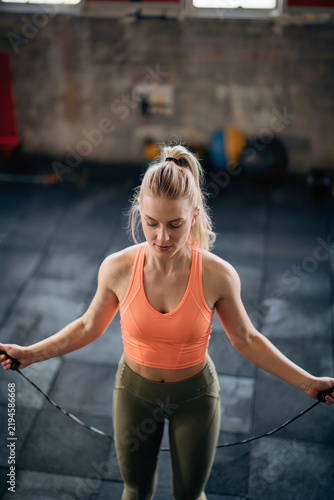 Focused athletic woman jump roping in a dim gym, wearing an orange cropped tank top and olive leggings while gripping the rope handles during an intense workout.