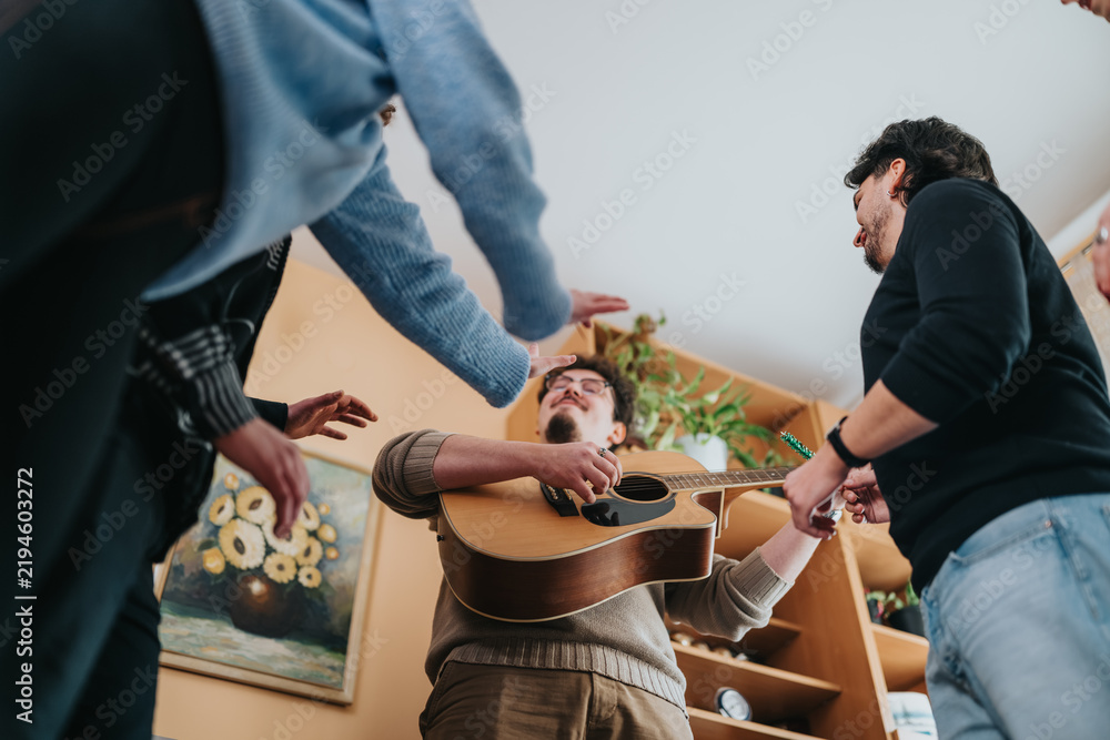 Fototapeta premium A young man plays an acoustic guitar while friends gather around him in a living room. The casual group sings and interacts during a relaxed home music session.
