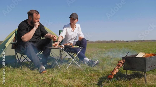 Young man and woman sitting in folding chairs, eating grilled meat and vegetables during a relaxing camping trip in a beautiful green meadow with a tent set up in the background