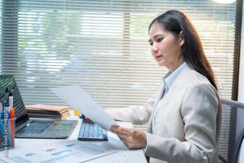 Determined businesswoman reviewing financial reports at contemporary office workspace with laptop and paperwork