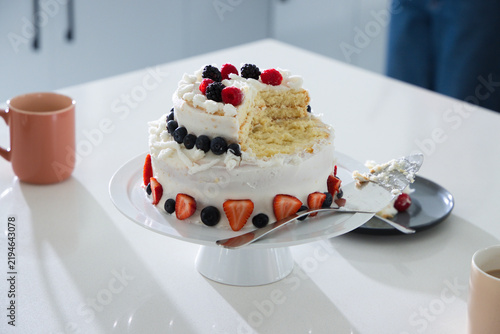 Two-tier white-frosted cake is sitting on pedestal stand on glossy counter, showing berry topping