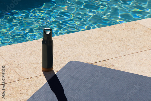 Matte gray water bottle is standing on travertine pool deck, casting long shadow near yoga mat