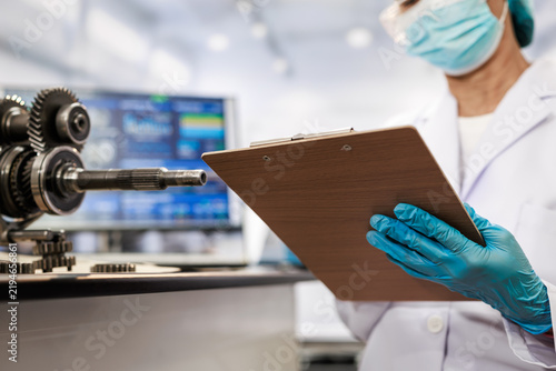 Masked female technician wearing blue gloves checks a clipboard beside industrial gears and a computer monitor in a clean modern laboratory.