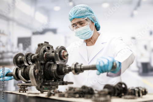Focused factory technician in protective clothing inspects a complex metal gear assembly on a workbench in a clean industrial workshop.