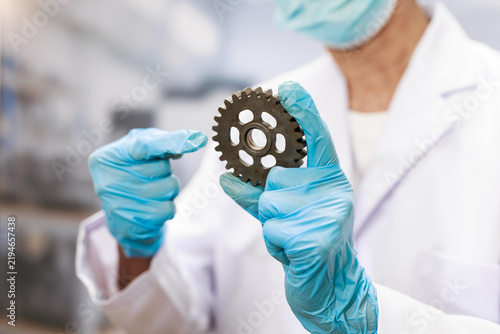 Scientist in a lab coat, mask and blue gloves holding a metal gear, inspecting an industrial part in a modern laboratory.