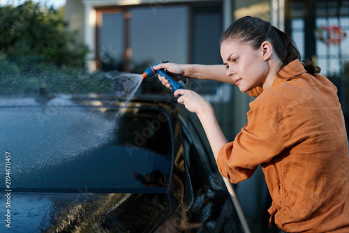 Konstfotografi A focused young woman in an orange shirt washes her dark car with a water hose spray nozzle outside her home