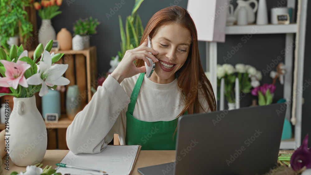 Obraz premium Woman florist holding smartphone to ear with hand on laptop and arranging flowers in a building; cheerful small business.