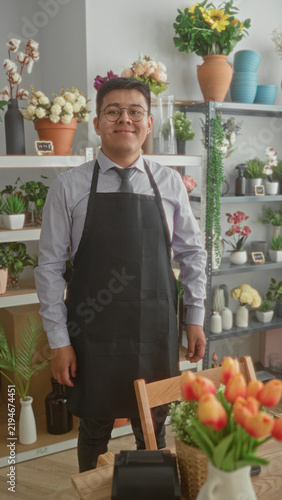 Fotografía Man florist wearing apron and tie with glasses, arms crossed and smiling in studio surrounded by potted flowers, bouquets and terracotta pots; friendly service
