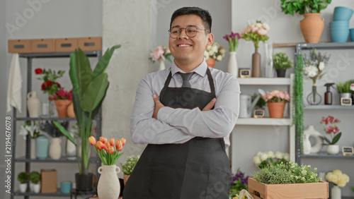 Photography Man florist wearing apron and tie holding an orange tulip bouquet, arms crossed and smiling inside a building with shelves of vases and potted plants; quiet pride