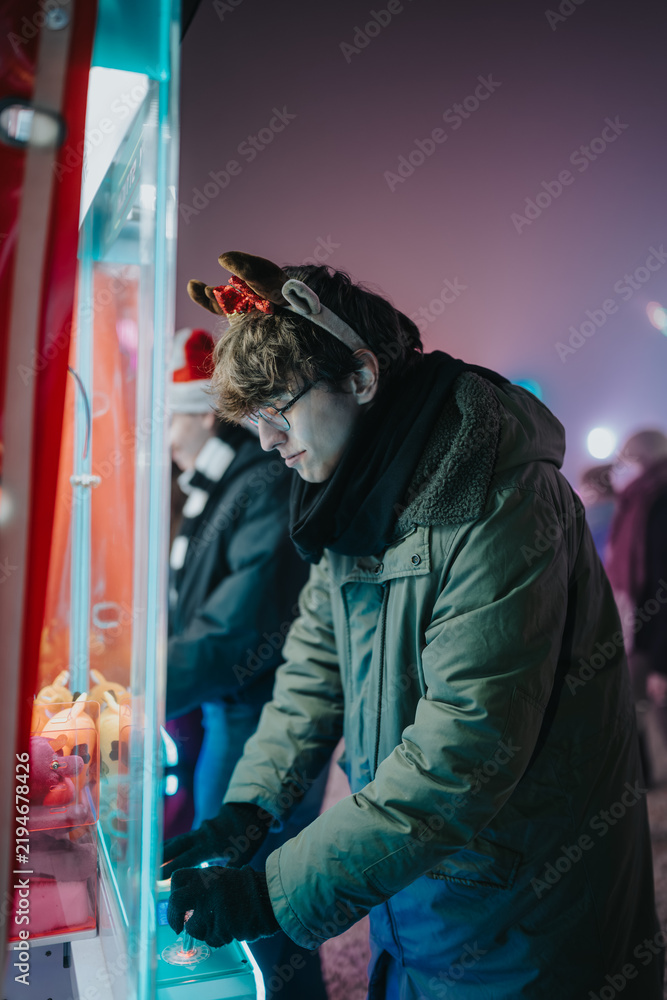 Fototapeta premium A young man in a parka and reindeer headband plays a claw crane game at a festive night market. The candid scene shows friends enjoying a neon-lit arcade booth during a cold holiday evening.