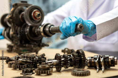 Obraz na plátně Mechanic in blue gloves holds a metal shaft beside a disassembled gearbox, with gears and machine parts spread across a workshop table