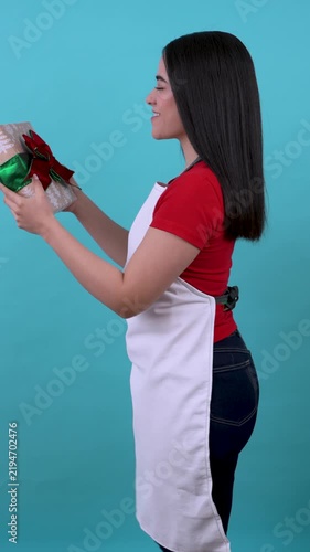Young latina woman wearing an apron joyfully presenting a wrapped christmas present on a blue background
