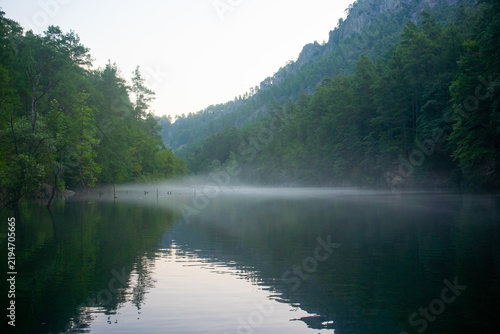 A symmetrical reflection on the calm surface of a lake nestled among forested hills in the early morning hours. A light mist and a serene atmosphere prevail.