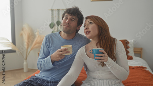 Man and woman sitting on bed holding blue and yellow coffee mugs, leaning toward each other and talking in a bright bedroom with pampas grass and window light; intimacy.