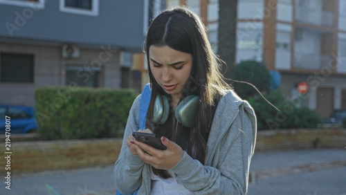 Obraz na płótnie Young woman with backpack and headphones holding smartphone, fingertip to temple, reading screen on street; concern