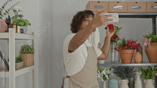 Obraz na plátně Man florist wearing apron holding smartphone, hand raised pointing to flowers in building; small business focus