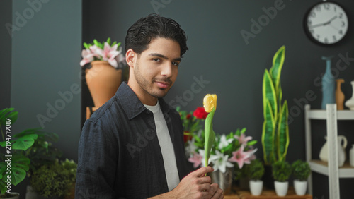 Canvas Print Man florist holding a yellow tulip in a plant filled studio with vases, pots and a wall clock; calm care