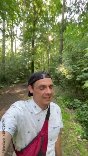 man walks along narrow dirt path in dense deciduous woods, sun rays glowing through leaves, warm shadows on ground, outdoor hiking mood today in peace calm