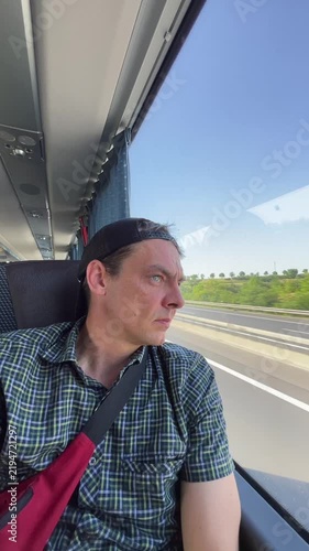 seated male passenger in plaid shirt and backward cap looks through large coach window at blurred fields and roadside greenery under clear blue sky sunlight man