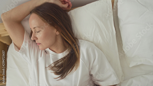 Cuadro en lienzo Woman lying in bed with arm above head and hand on stomach on white sheets beside a rattan nightstand in building; serenity rest