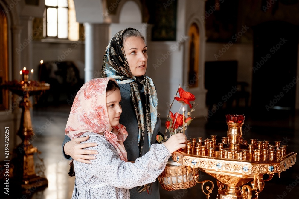 Obraz premium mom and daughter put candles and pray in the Orthodox church, congratulating each other on Easter, bright Easter in the temple, mom and daughter cross themselves looking at the icon