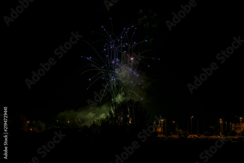 Cuadro en lienzo Blue fireworks sparks and trails rising above tree silhouette at night