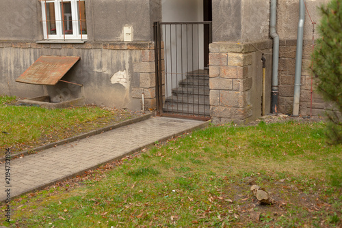 Grassy approach path leading to gated stair and stoop, worn pavement edging with scattered leaves and moss, small front yard and muted tones conveying peaceful residential arrival 