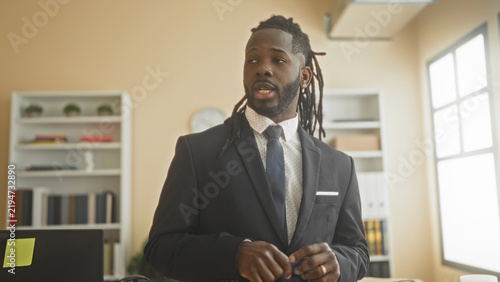 Картината върху платно Man in suit with dreadlocks holding a pen and gesturing with hands while speaking at a desk near bookshelf and window in office; confidence leadership