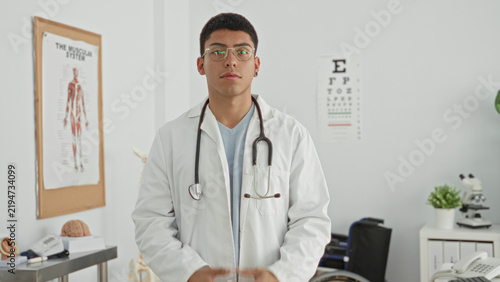Fotografie Man doctor in white coat adjusts stethoscope while holding collar in clinic exam room with eye chart and anatomy poster visible; professional calm