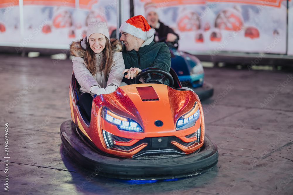 Obraz premium A young couple rides together in a bright orange bumper car, smiling and laughing. They enjoy a festive amusement park ride at a winter fair with holiday decorations.