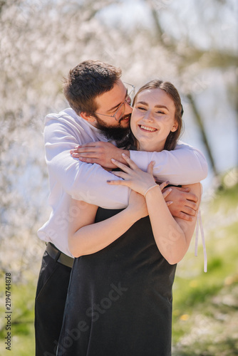 Картината върху платно Young couple embracing outdoors near a body of water, surrounded by blooming tre