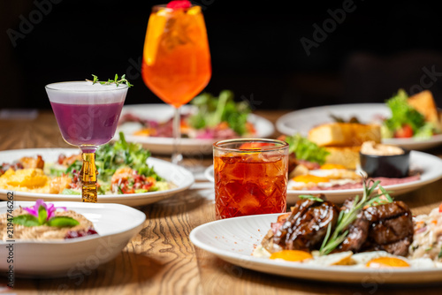 An elegant table spread featuring gourmet dishes, a grilled steak with rosemary, and colorful cocktails including a purple sour and orange spritz in a dimly lit restaurant setting.