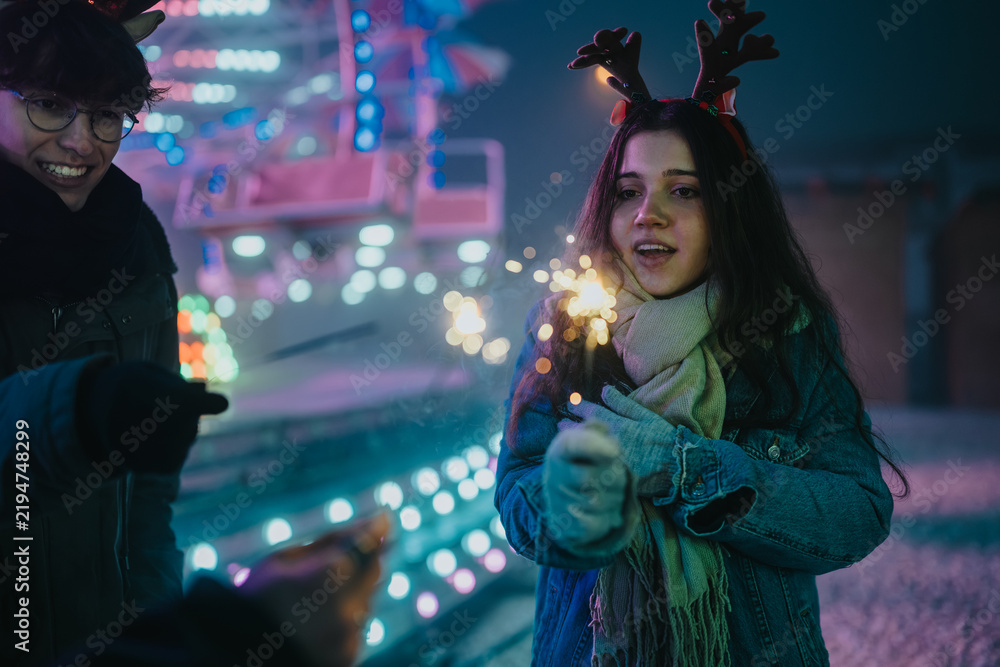 Obraz premium Two friends hold sparklers while enjoying a festive winter night. The scene shows a young pair celebrating among colorful lights at a holiday market.