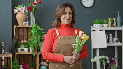 фотографія Woman florist in apron cradles yellow and purple tulips, smiles and tilts head while holding bouquet beside shelf and clock in studio; warmth care