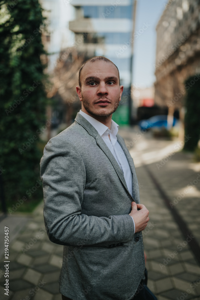 Fototapeta premium A young professional man wearing a grey blazer looks confidently to the side for a business portrait. The urban street setting provides a casual outdoor professional atmosphere.