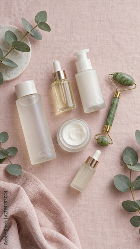 Top view flat lay of unlabeled skincare bottles, a jade roller and eucalyptus on pink linen