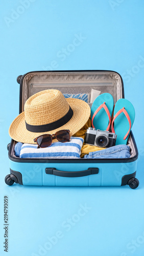 Open suitcase with a straw hat, sunglasses, flip flops and a camera on sky blue