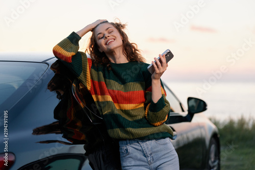Принти на полотні Happy young woman in striped sweater leaning on car at sunset, holding smartphone by the sea
