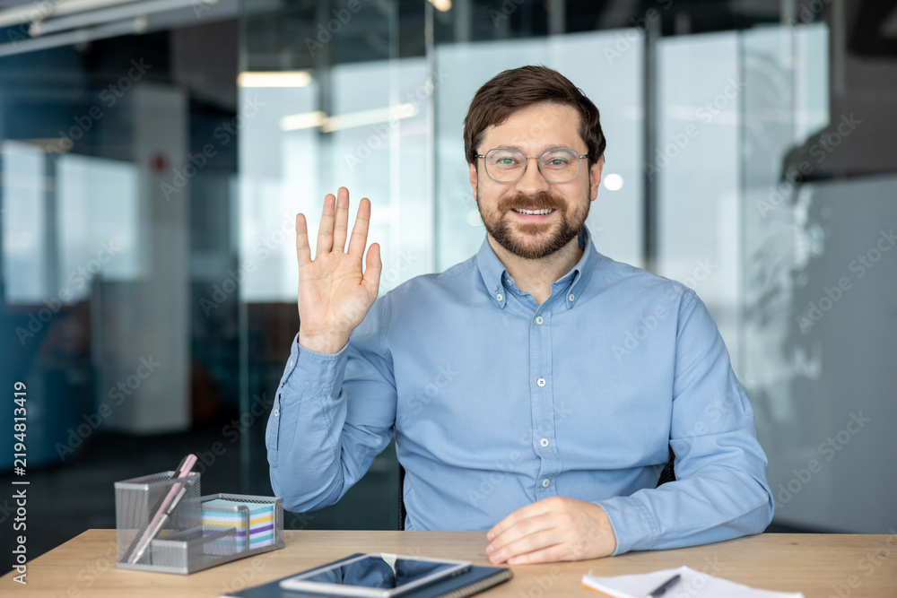 Fototapeta premium A portrait of a smiling young man in an office; he is sitting at his desk, waving hello, and starting a remote conversation on camera