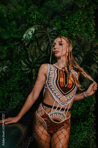 Female nightclub performer poses in patterned festival stagewear and fishnet tights beside a lush green tropical wall, creating a vibrant image for nightlife and fashion promotion