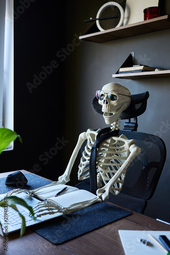 Foto Skeleton wearing glasses and tie sitting at office desk with laptop, notebook an
